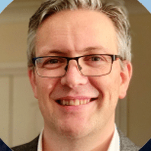 A man with short grey hair and glasses smiles at the camera. He is indoors, and the background is softly blurred.