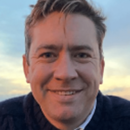 A person with short, light brown hair smiles outdoors with a sunset sky in the background, wearing a dark jumper over a collared shirt.