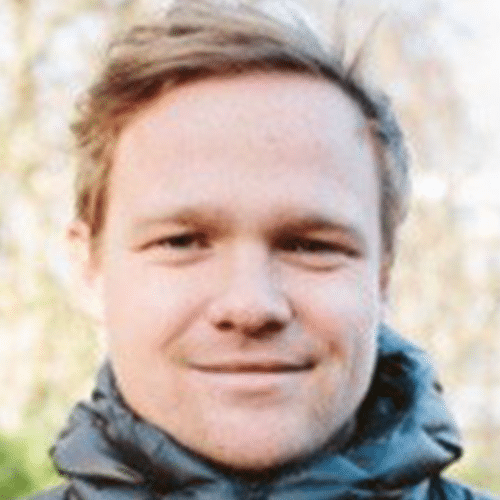 A person with light skin and short, light brown hair is outdoors, wearing a grey puffy jacket. The background is blurred with hints of trees and sky. The person is smiling slightly.