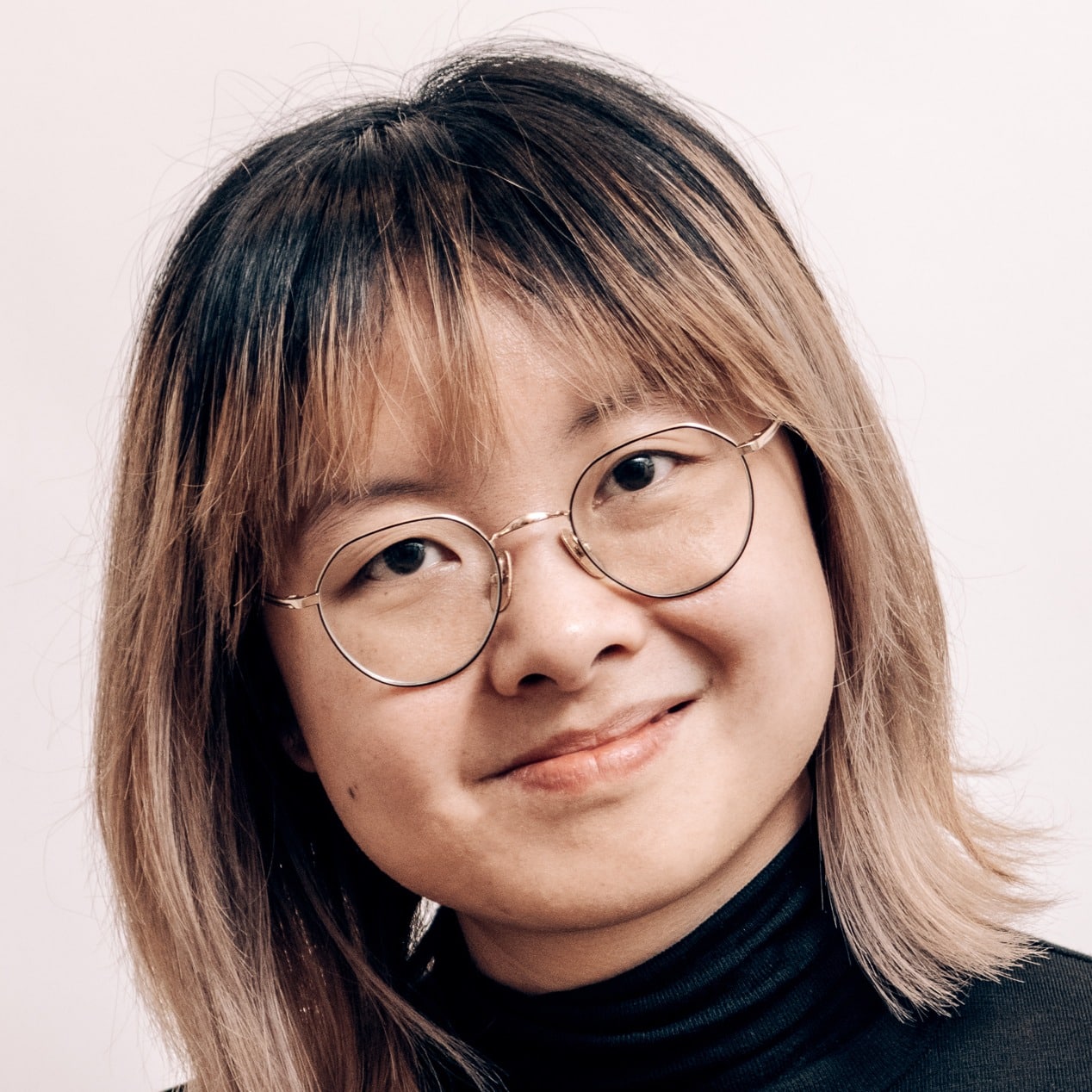 Person with shoulder-length, light brown hair, round glasses, and a black polo neck smiles gently against a plain background.