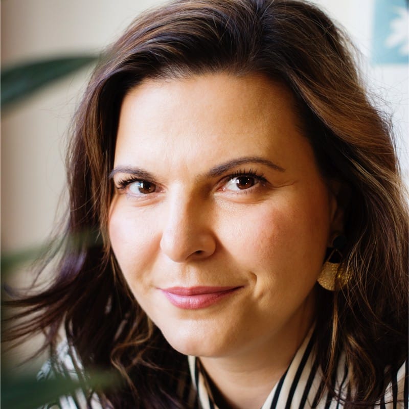 A woman with long brown hair and light skin smiles slightly, looking at the camera. She wears gold earrings and a black and white striped top. There is a blurred green leaf in the foreground.