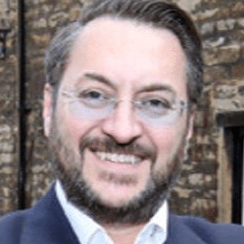 A man with glasses and a beard smiles at the camera, standing in front of a brick wall.