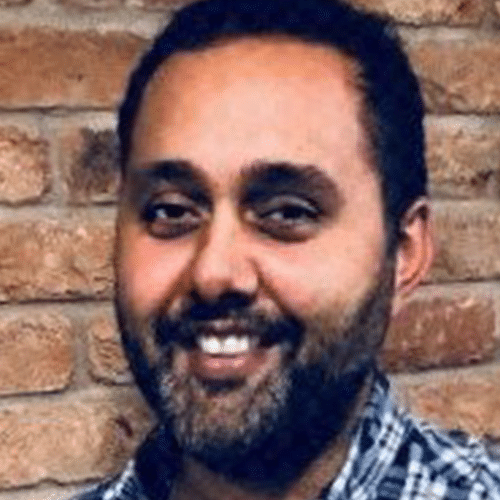 A man with a beard and moustache, wearing a checked shirt, smiles in front of a brick wall.