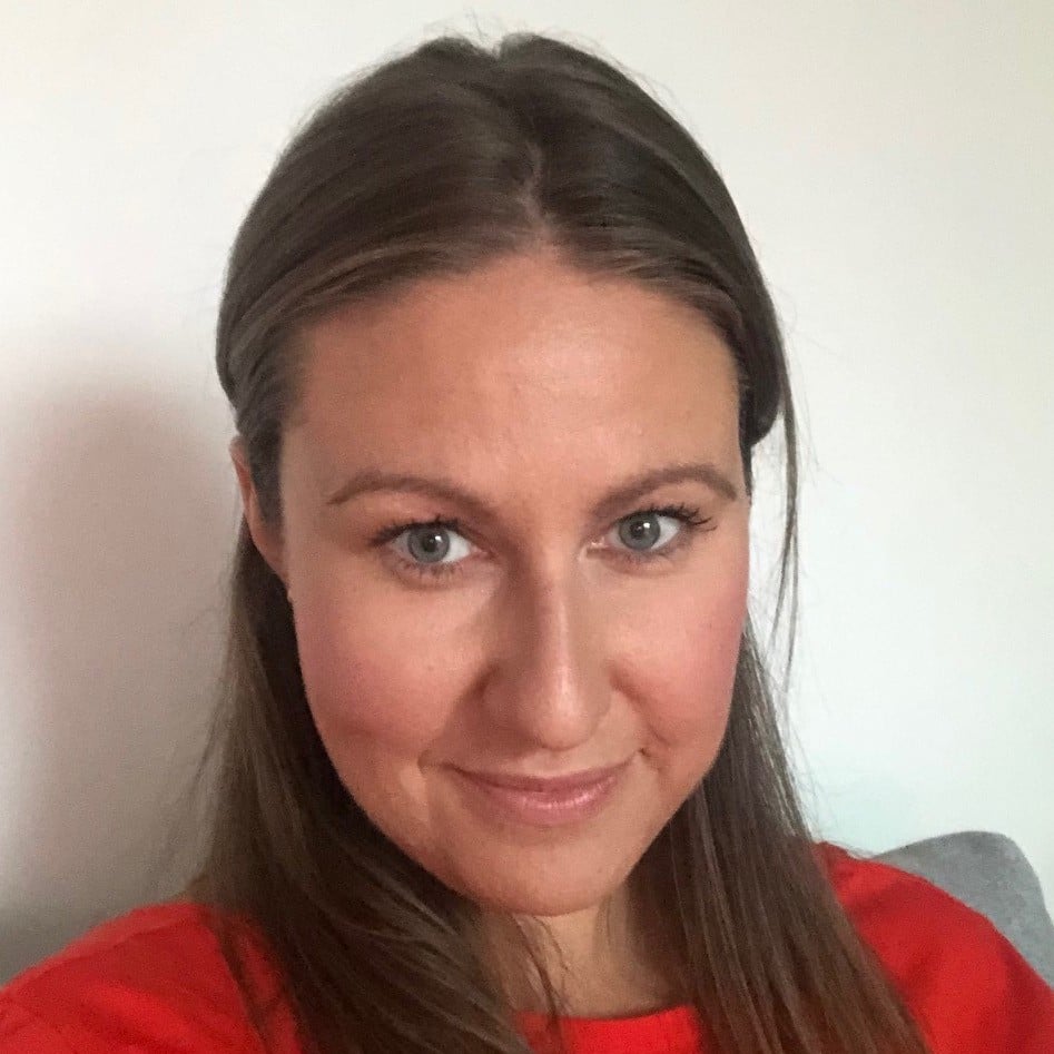 A woman with long brown hair and blue eyes, wearing a red jumper, smiles slightly at the camera in front of a plain light-coloured background.