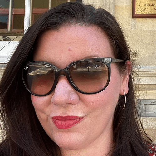 Woman with long dark hair wearing large black sunglasses, hoop earrings, and red lipstick, standing outside in front of a light-coloured stone building with a plaque and window in the background.
