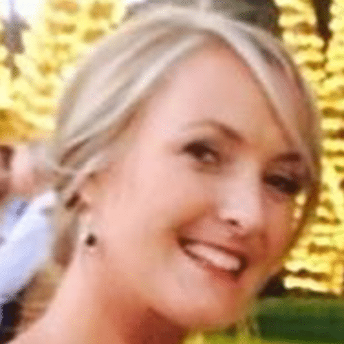 Smiling woman with blonde hair poses outdoors in front of blurred yellow lights and greenery.