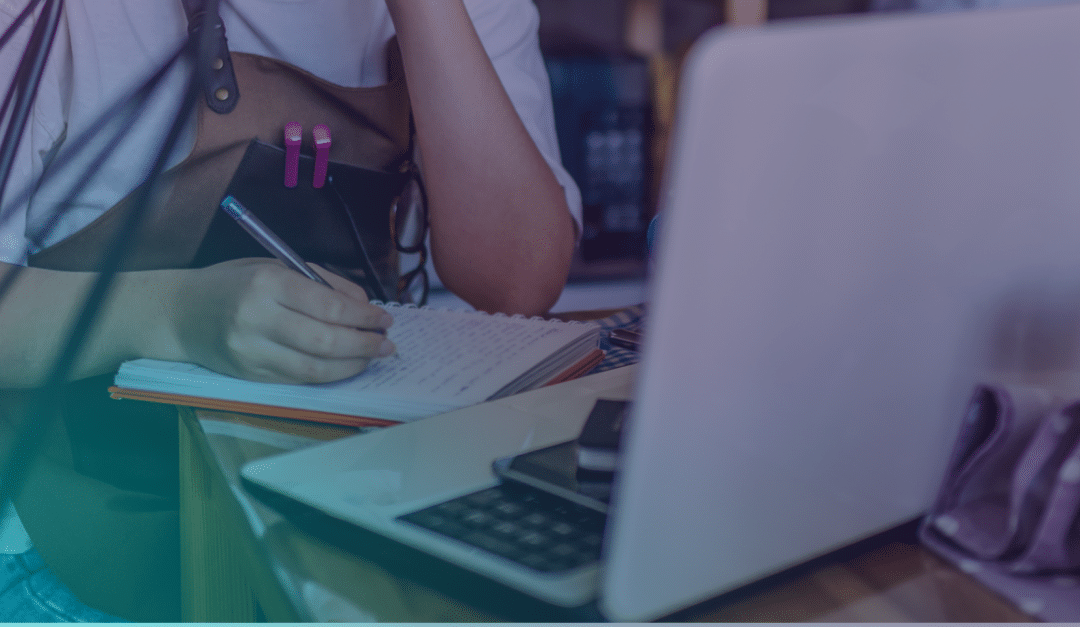 A person wearing an apron writes in a notebook with a pen, sitting at a table with a laptop, calculator, and mobile.
