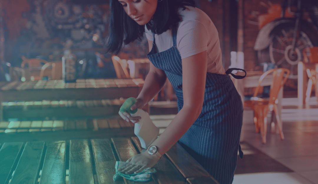 A woman wearing an apron cleans a wooden table with a spray bottle and cloth in a dimly lit restaurant.
