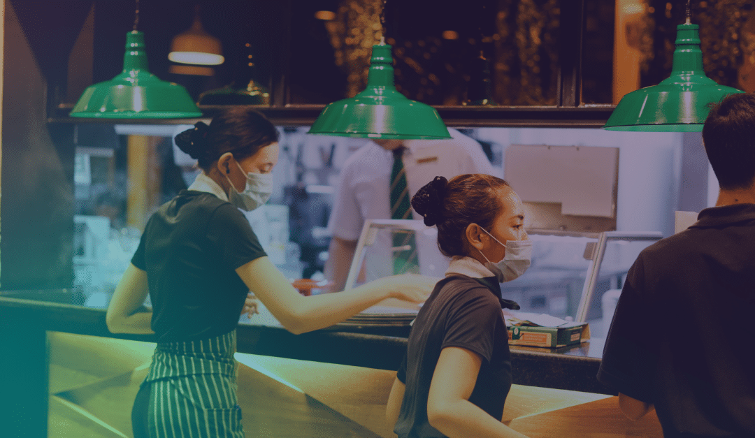 Two food service workers wearing masks stand behind a counter under green lamps, preparing food. Another person is visible in the background, also wearing a mask, in a restaurant or café setting.