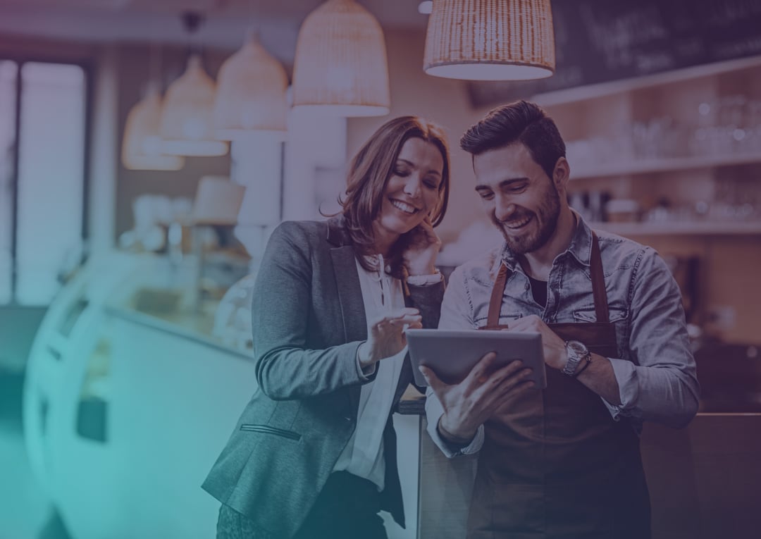 Two people, one in business attire and one in an apron, smile whilst looking at a tablet together inside a cosy café. Warm lighting and a display case are visible in the background.