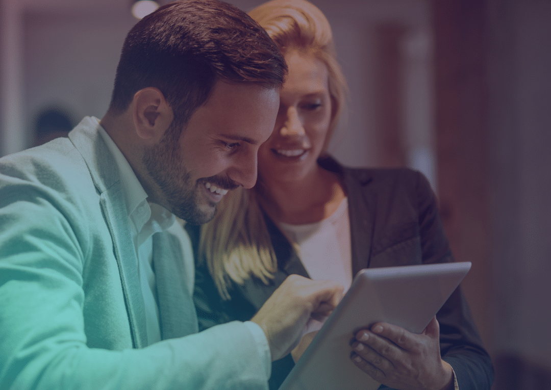 Two people in business attire smile while looking at and using a tablet together in an office setting.