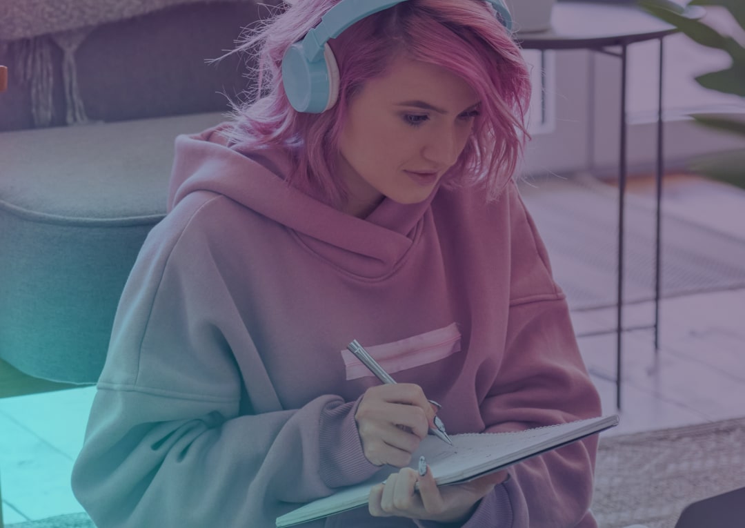 A person with pink hair and blue headphones wears a pink hoodie and writes in a notebook whilst sitting on the floor in a cosy indoor setting.