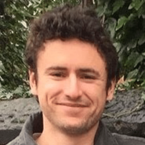 A young man with curly brown hair and a slight beard smiles outdoors, with green foliage in the background.
