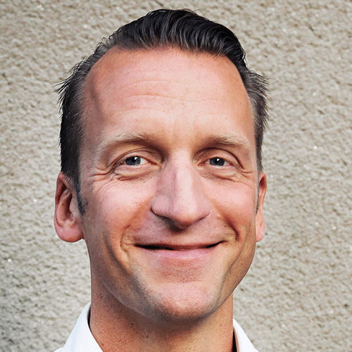 A man with short, neatly styled hair smiles at the camera in front of a textured beige wall.