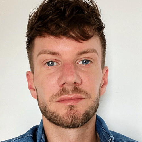 A man with short brown hair, blue eyes, and a trimmed beard is facing the camera against a plain white background. He is wearing a blue shirt.