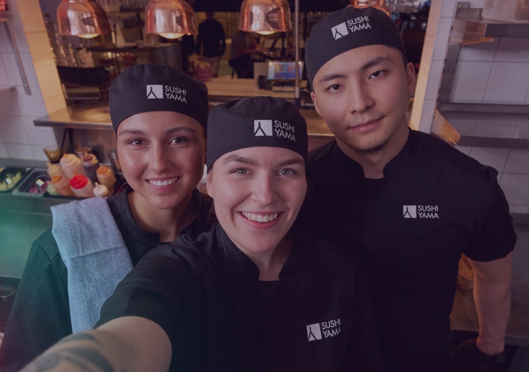 Three smiling people in Sushi Yama uniforms and hats stand closely together in a restaurant kitchen, posing for a selfie.