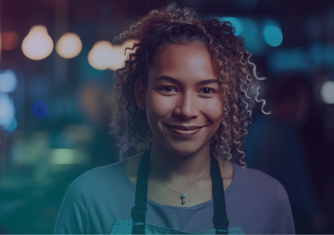 A young woman with curly hair, wearing an apron and a necklace, smiles softly in a warmly lit indoor setting with blurred lights in the background.