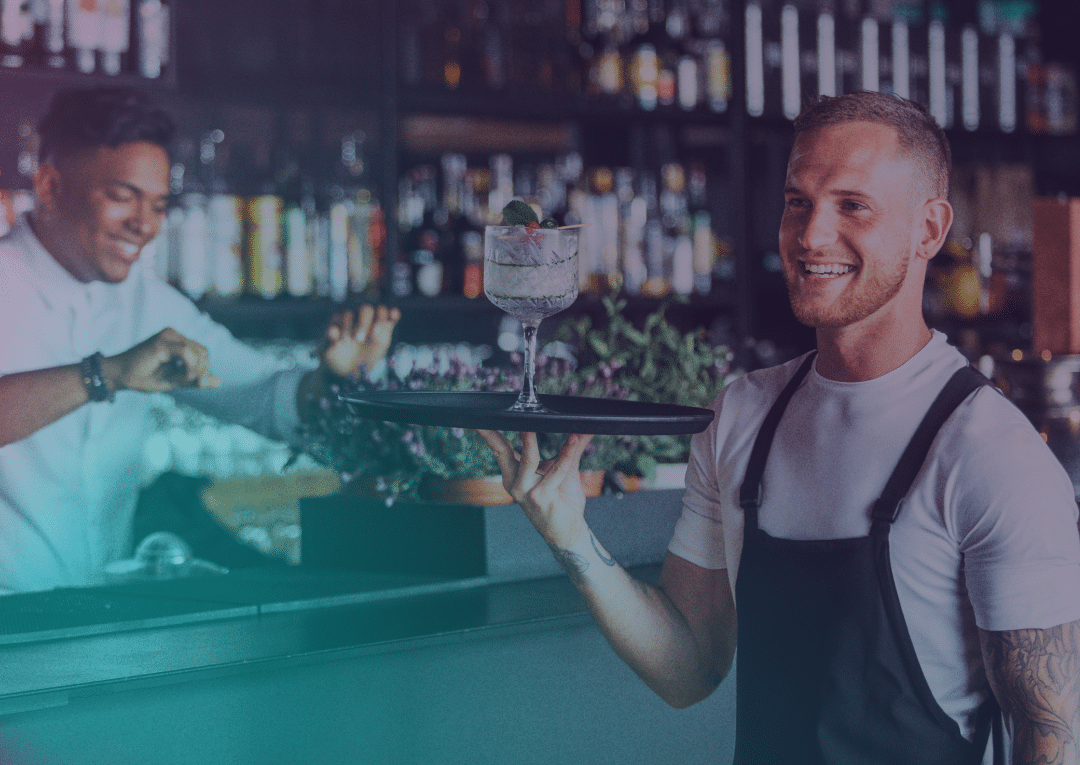 A smiling waiter in a black apron holds a cocktail on a tray, whilst a barman prepares drinks behind a bar filled with bottles.
