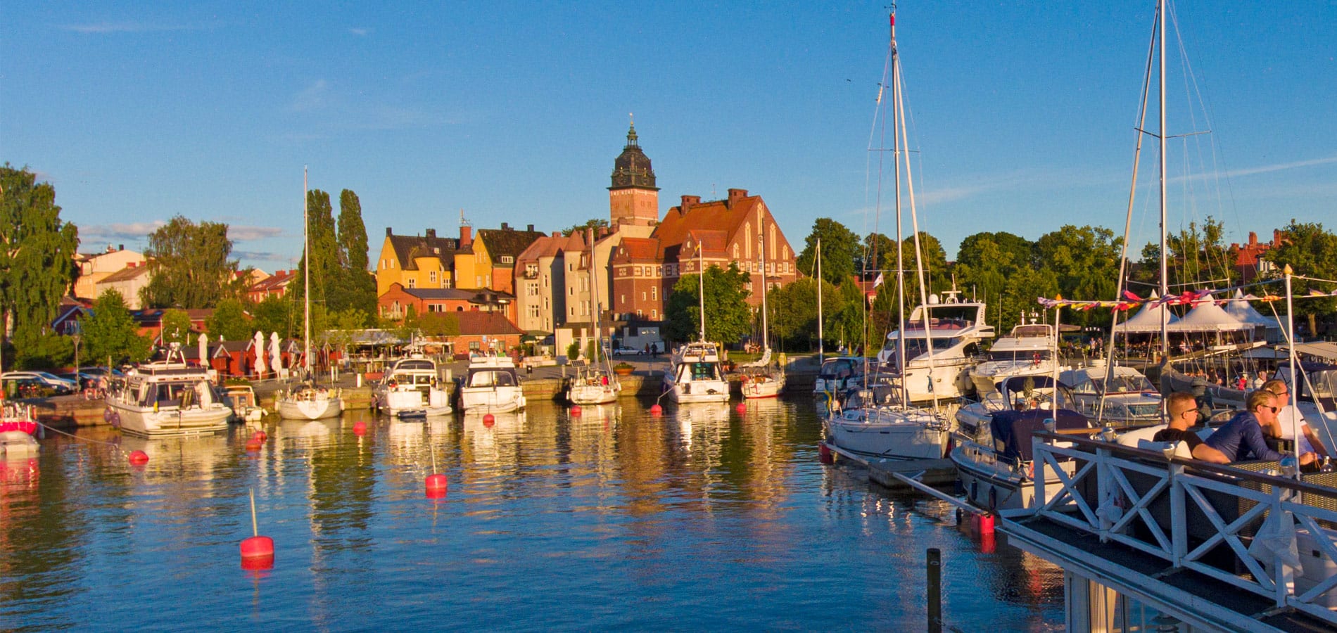 Boats moored at a marina with colourful historic buildings and a church tower in the background, under a clear blue sky, with people sitting on a waterside deck.
