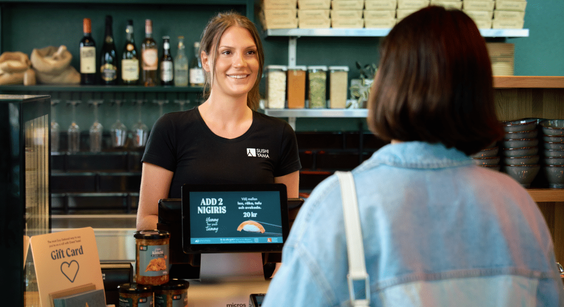 A smiling cashier stands behind a counter in a modern restaurant, engaging with a customer. Various products, wine bottles, and a gift card display are visible on the counter and shelves.