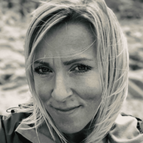 A woman with fair hair smiles slightly at the camera outdoors. She is wearing a jacket and the background appears blurred, possibly showing a natural setting. The image is in black and white.