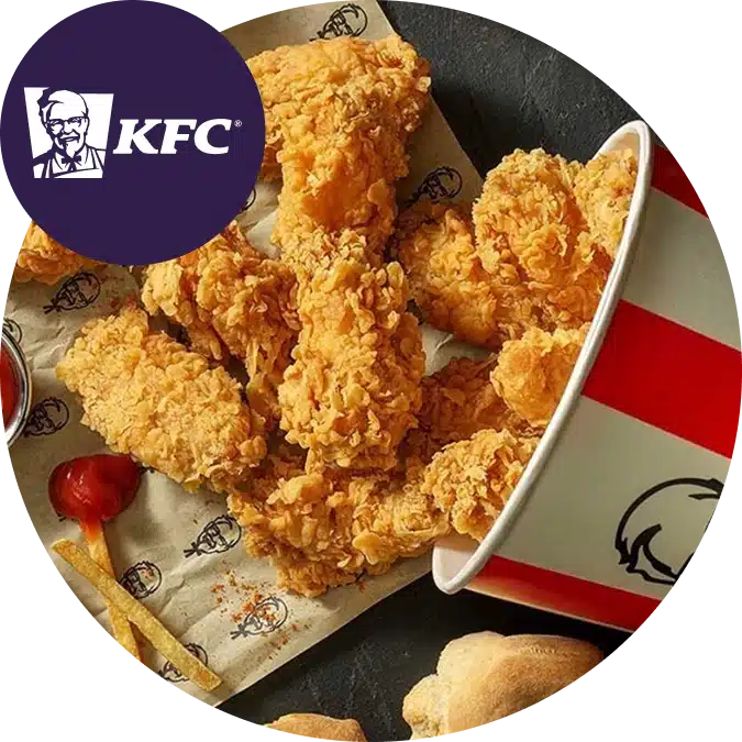 KFC fried chicken strips spill from a red and white KFC bucket onto branded paper, with chips, a scone, and tomato sauce on the side. The KFC logo is in the top left corner.