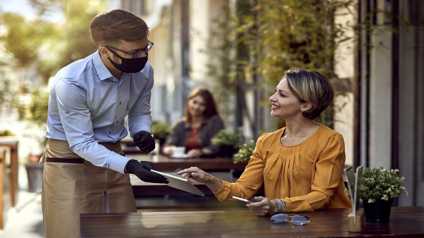 A masked waiter hands a bill to a smiling woman seated at an outdoor café table. She holds a card and mobile. Another person sits at a table in the background. There are small potted plants on the tables.