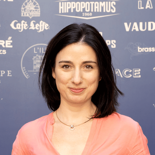 Une femme aux cheveux foncés mi-longs, vêtue d'un chemisier rose pâle et portant un collier, se tient debout, souriante, devant un fond bleu orné de logos et de texte blancs.