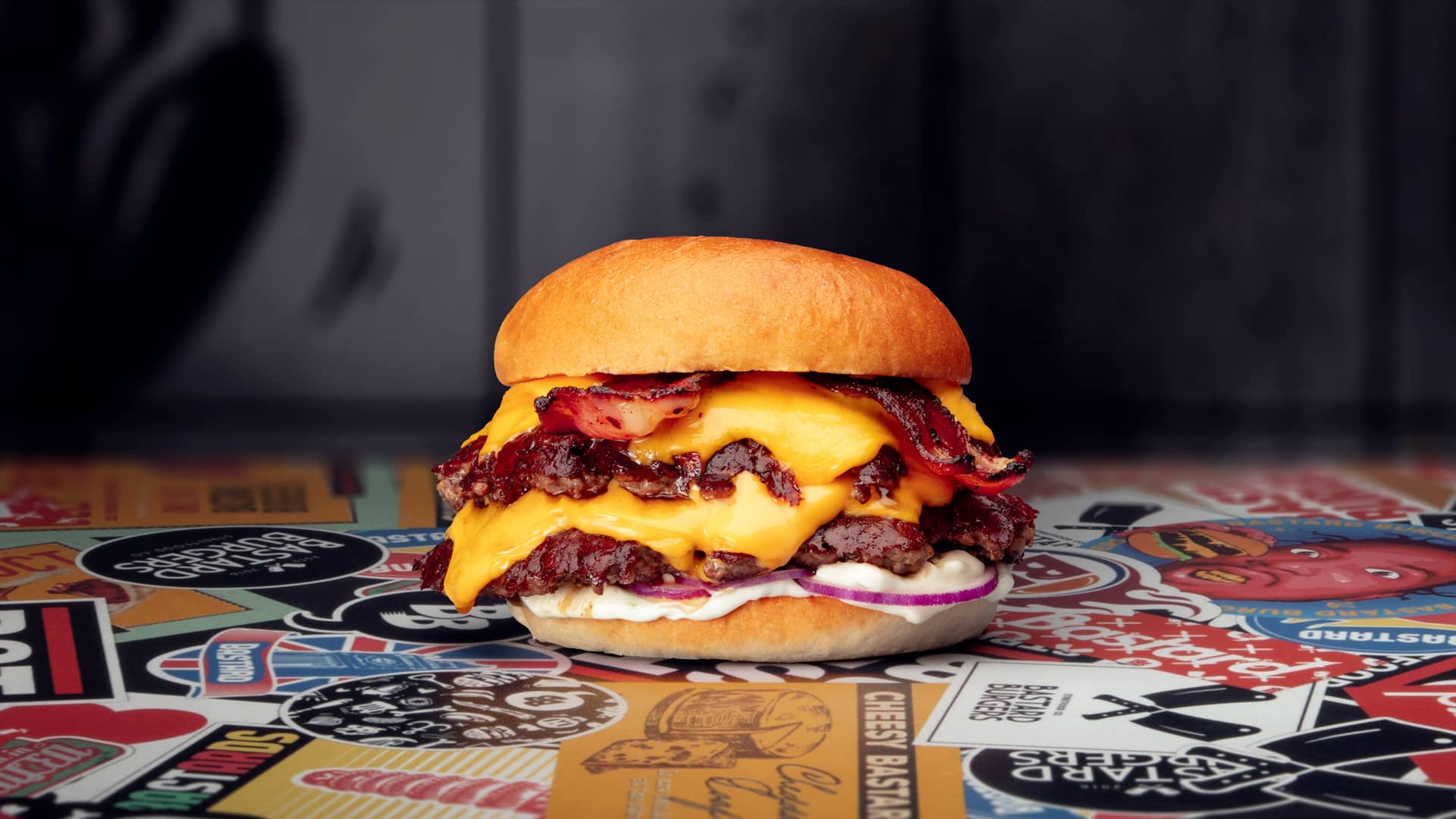 Un cheeseburger composé de deux steaks hachés, de fromage cheddar fondu, de bacon croustillant, de mayonnaise et d'oignons, le tout dans un petit pain, posé sur une surface colorée recouverte d'autocollants.