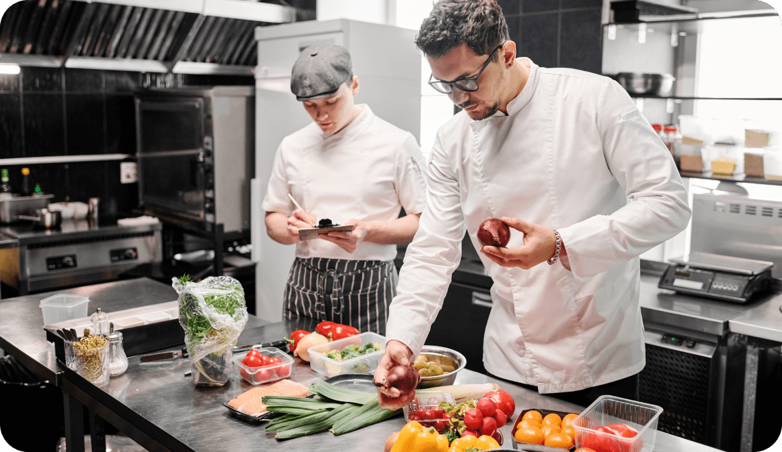 Dans une cuisine professionnelle, deux chefs préparent des légumes frais sur un plan de travail en inox ; l'un d'eux dispose les légumes tandis que l'autre prend des notes sur un bloc-notes à pince. Divers légumes et ingrédients sont étalés sur le plan de travail.