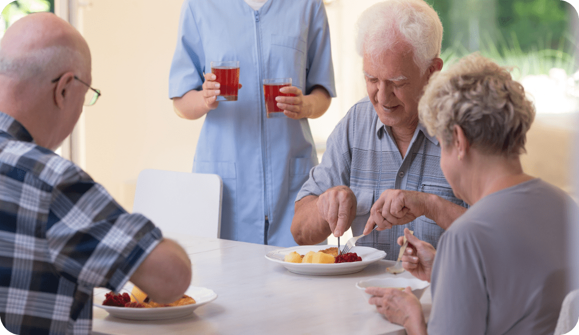 Trois personnes âgées sont assises à une table à manger, tandis qu'un soignant en uniforme bleu se tient debout à l'arrière-plan, tenant deux verres de jus de fruits rouge. L'ambiance semble joyeuse et conviviale.
