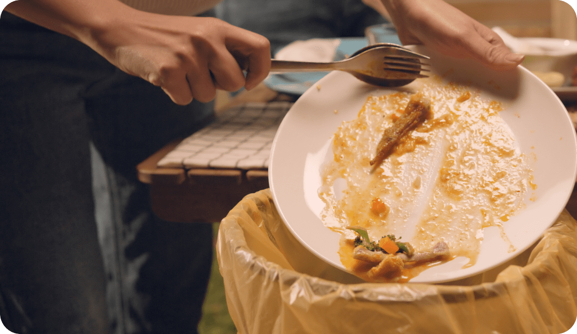 Une personne utilise une fourchette pour racler les restes d'un plat blanc et les jeter dans une poubelle jaune, afin d'éliminer les déchets alimentaires après le repas.