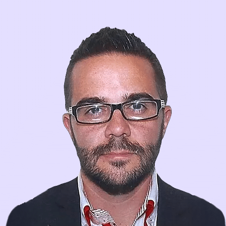 Un homme aux cheveux bruns courts, portant une barbe et une moustache, avec des lunettes rectangulaires, une veste foncée et une chemise à col ornée de touches rouges et blanches, sur un fond violet clair uni.