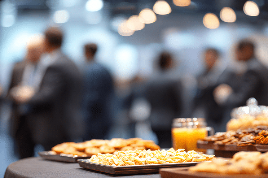 Lors d'un événement de réseautage, l'attention se porte sur une table garnie de viennoiseries et de jus d'orange, tandis qu'à l'arrière-plan, flou et baigné d'une lumière tamisée, des groupes de personnes en tenue professionnelle discutent.