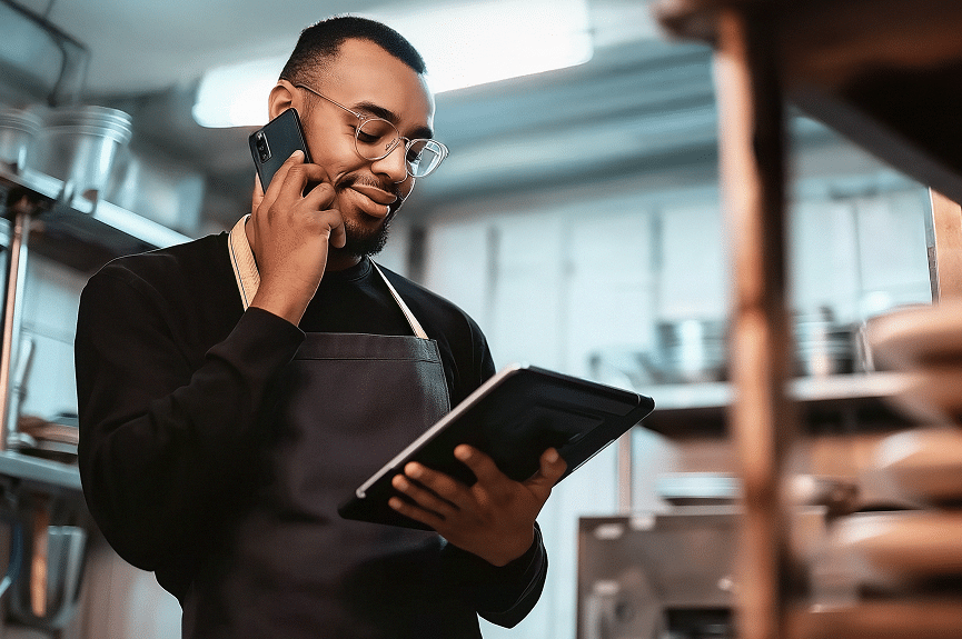 Un homme portant des lunettes et un tablier se tient debout dans une cuisine ; il parle au téléphone tout en regardant une tablette, entouré d'étagères et d'ustensiles de cuisine.