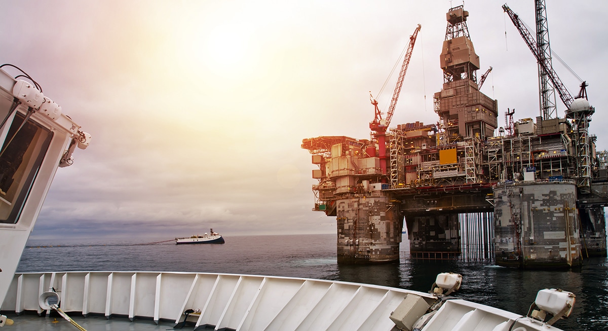 Une plate-forme pétrolière offshore se dresse dans l'océan sous un ciel radieux, avec des grues sur son pont. Un navire s'approche de la plate-forme, et la scène est filmée depuis un autre navire.