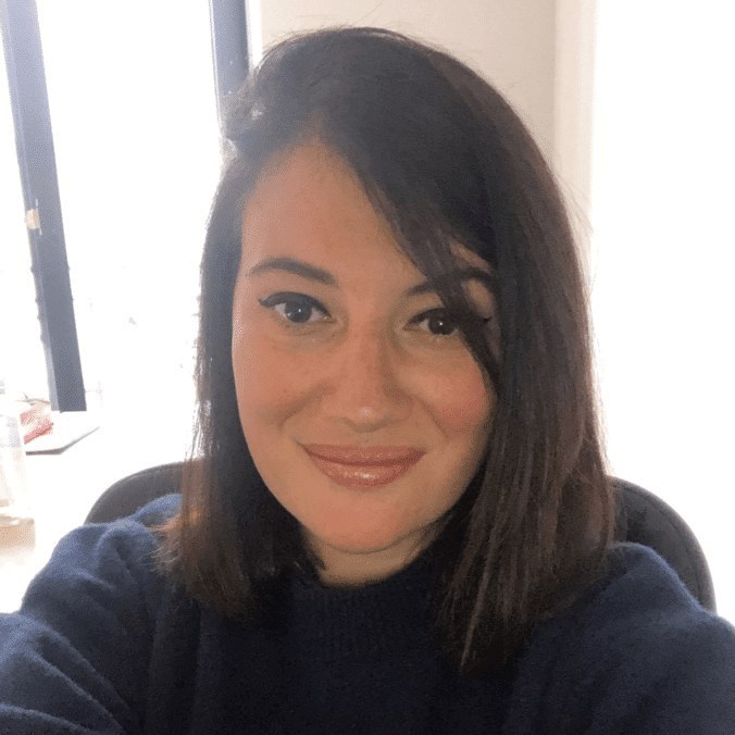 Une femme aux cheveux bruns raides tombant jusqu'aux épaules, les yeux soulignés d'un trait d'eyeliner en aile, sourit à la caméra à l'intérieur, vêtue d'un pull foncé. La lumière pénètre par une fenêtre toute proche.
