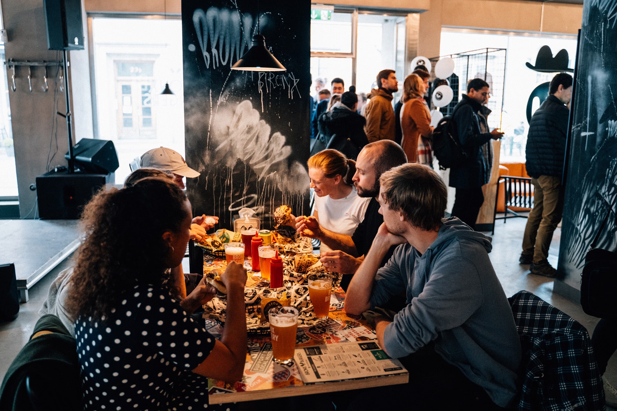 Dans un restaurant urbain animé, un groupe de six personnes est assis autour d'une table sur laquelle sont disposés des plats, des boissons et des condiments. On aperçoit d'autres clients et des œuvres d'art de style graffiti en arrière-plan.