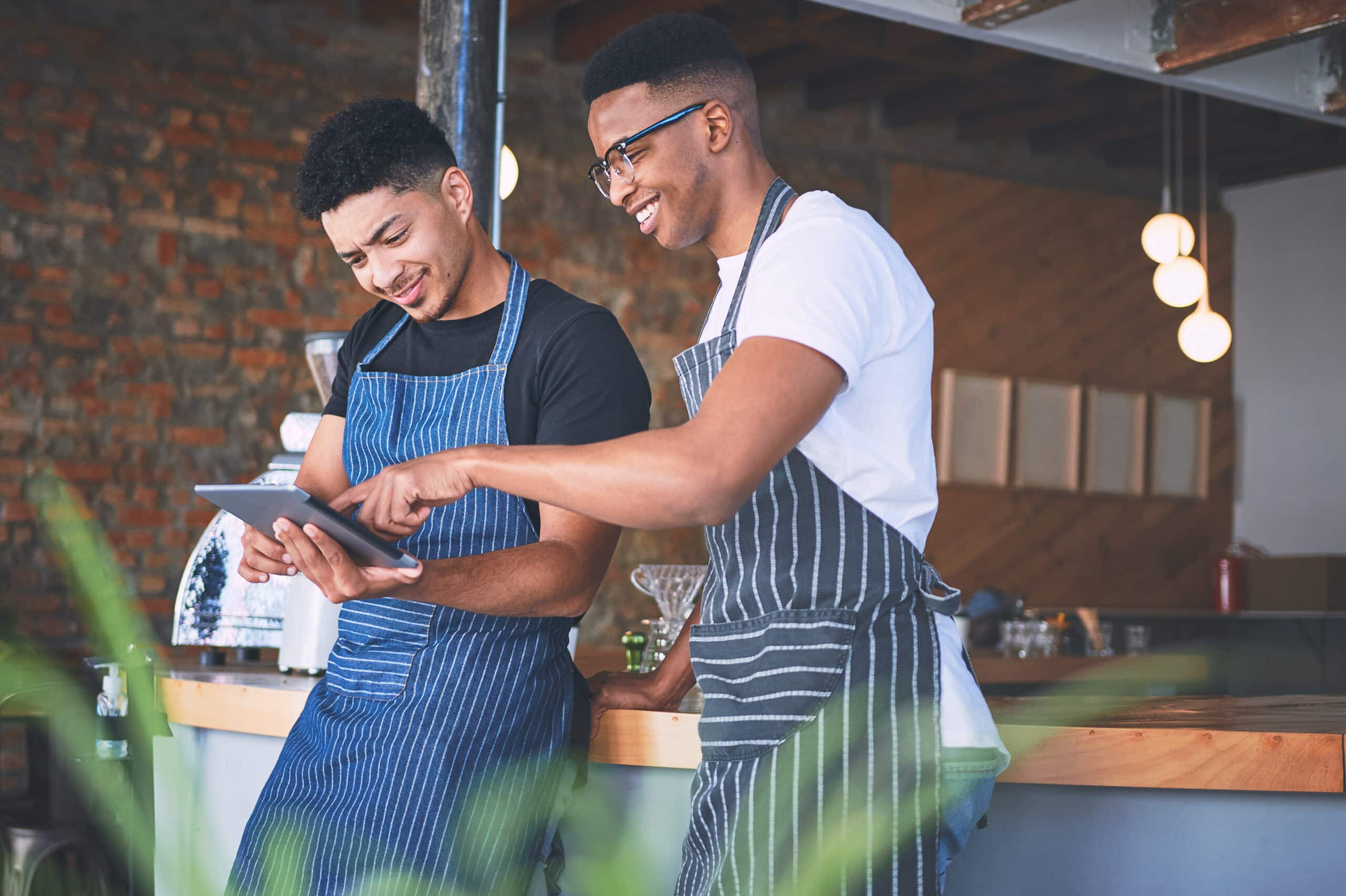 Deux jeunes hommes, vêtus de tabliers, se tiennent derrière le comptoir d'un café moderne ; ils sourient et regardent ensemble une tablette.