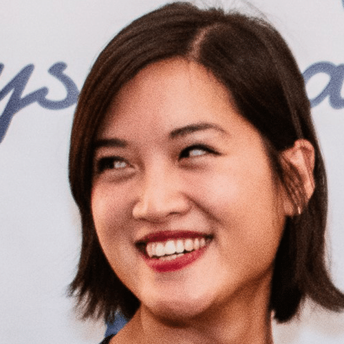 Une femme aux cheveux courts et foncés sourit en regardant vers sa droite. À l'arrière-plan, on distingue partiellement un texte bleu sur fond blanc.