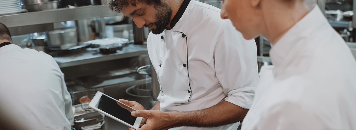 Deux chefs dans une cuisine professionnelle : l'un utilise une tablette tandis que l'autre l'observe. On aperçoit du matériel et des ustensiles de cuisine à l'arrière-plan.