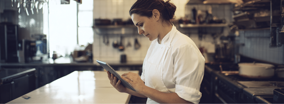 Un chef en tenue blanche se tient debout dans une cuisine professionnelle ; il consulte et utilise une tablette, tandis qu'on aperçoit en arrière-plan des étagères, des casseroles et du matériel de cuisine.