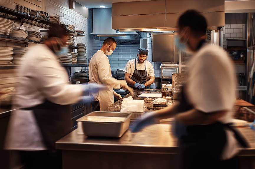 Des chefs en uniforme et portant des masques travaillent ensemble dans la cuisine animée et moderne d'un restaurant, préparant des plats sur un plan de travail en inox entouré d'étagères remplies d'assiettes. La scène dégage une impression de dynamisme et de travail d'équipe.