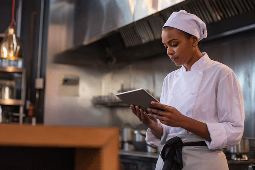 Un chef, vêtu d'un uniforme blanc et d'un foulard, se tient debout dans une cuisine professionnelle, les yeux rivés sur une tablette. On aperçoit à l'arrière-plan des appareils électroménagers et des ustensiles de cuisine en inox.