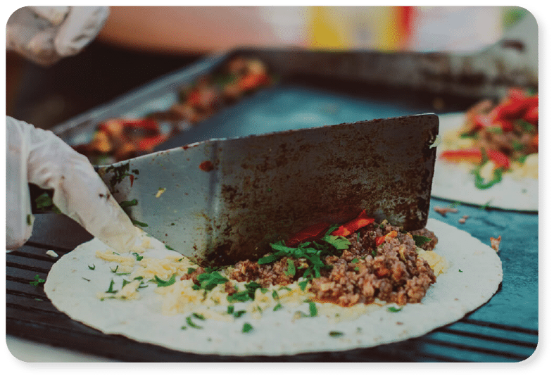 Une main gantée tient une spatule et fait cuire sur une plaque chauffante une tortilla garnie de viande hachée, de fromage, d'herbes aromatiques et de poivrons rouges.