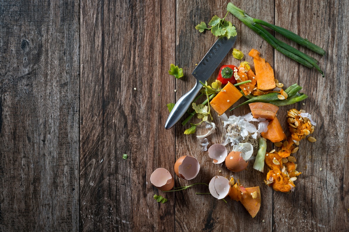 Des restes de légumes, des coquilles d'œufs et un couteau sont posés sur un plan de travail rustique en bois. Des oignons verts, de la coriandre et des morceaux de légumes orange sont éparpillés parmi les déchets alimentaires.