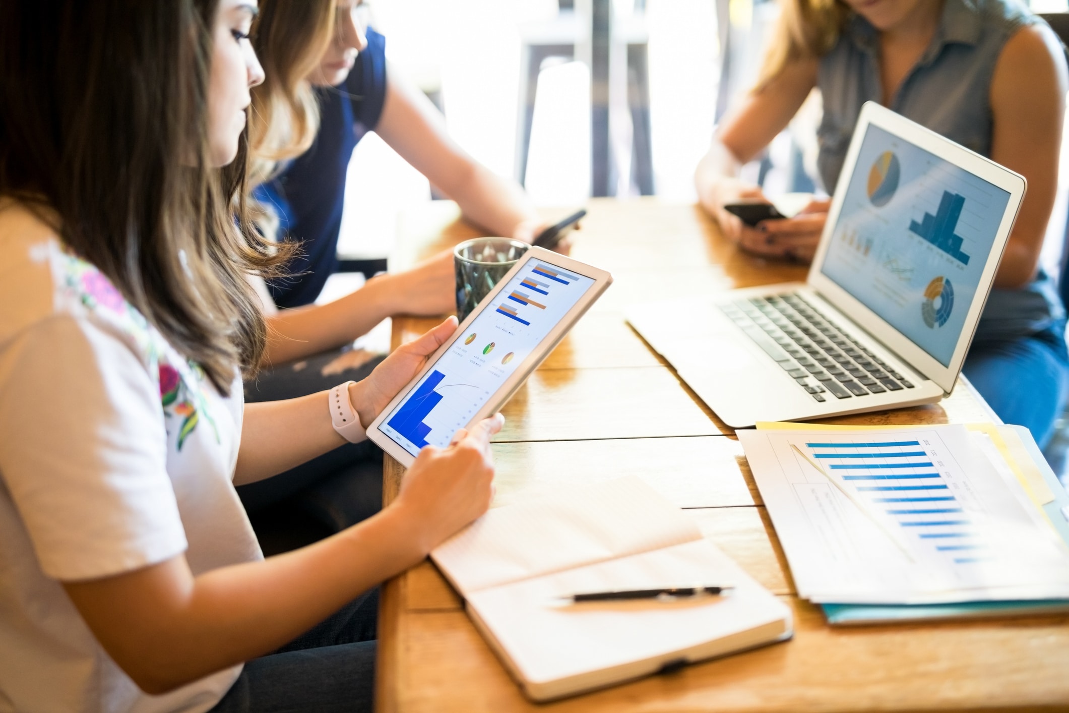 Trois femmes sont assises autour d'une table en bois sur laquelle se trouvent un ordinateur portable affichant des tableaux, une tablette présentant des graphiques, des cahiers et des téléphones. Elles semblent travailler ensemble ou analyser des données.