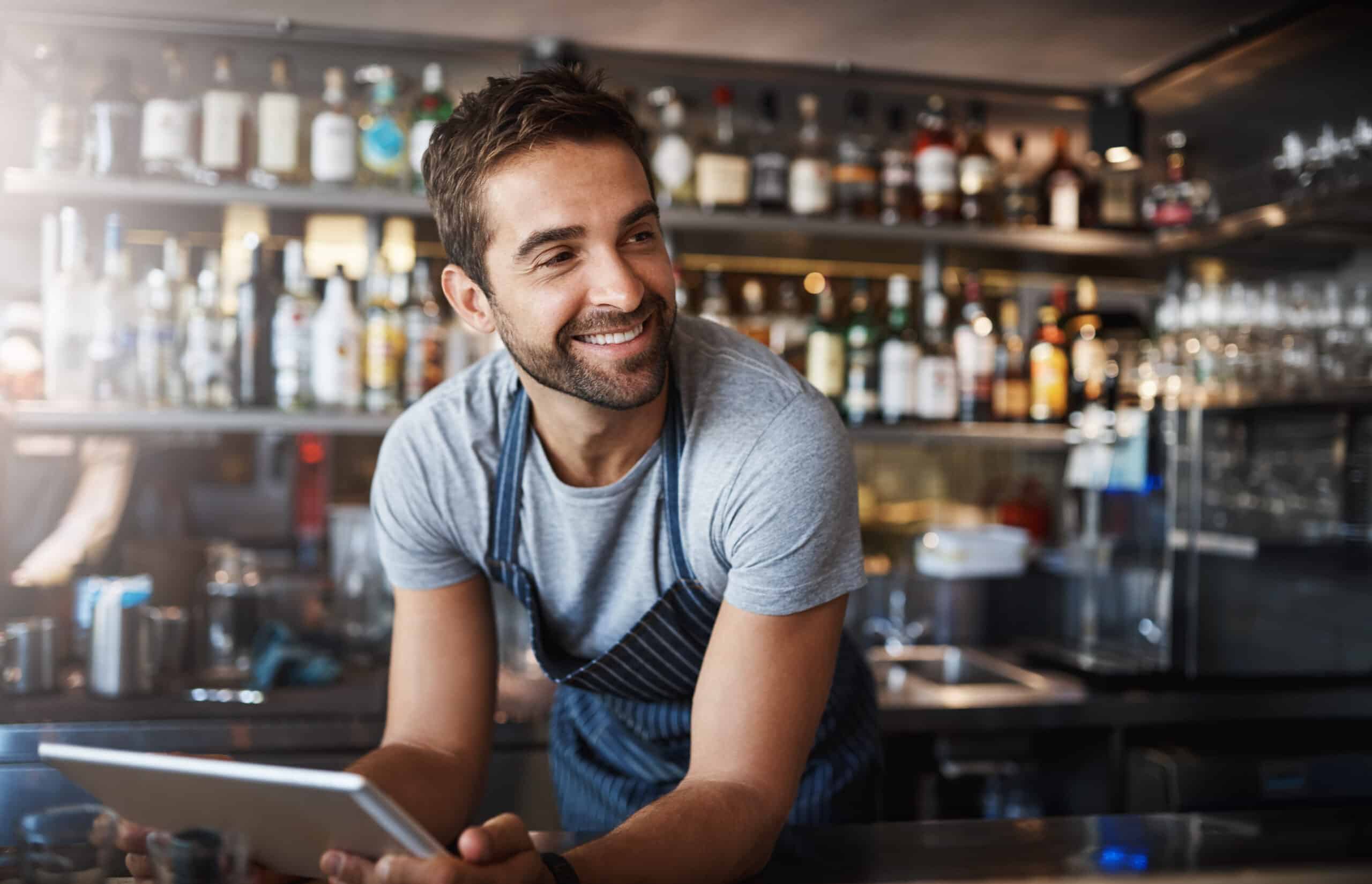 Un homme souriant, vêtu d'un t-shirt gris et d'un tablier, se tient derrière un comptoir, une tablette à la main. On aperçoit des bouteilles et des verres sur les étagères en arrière-plan.