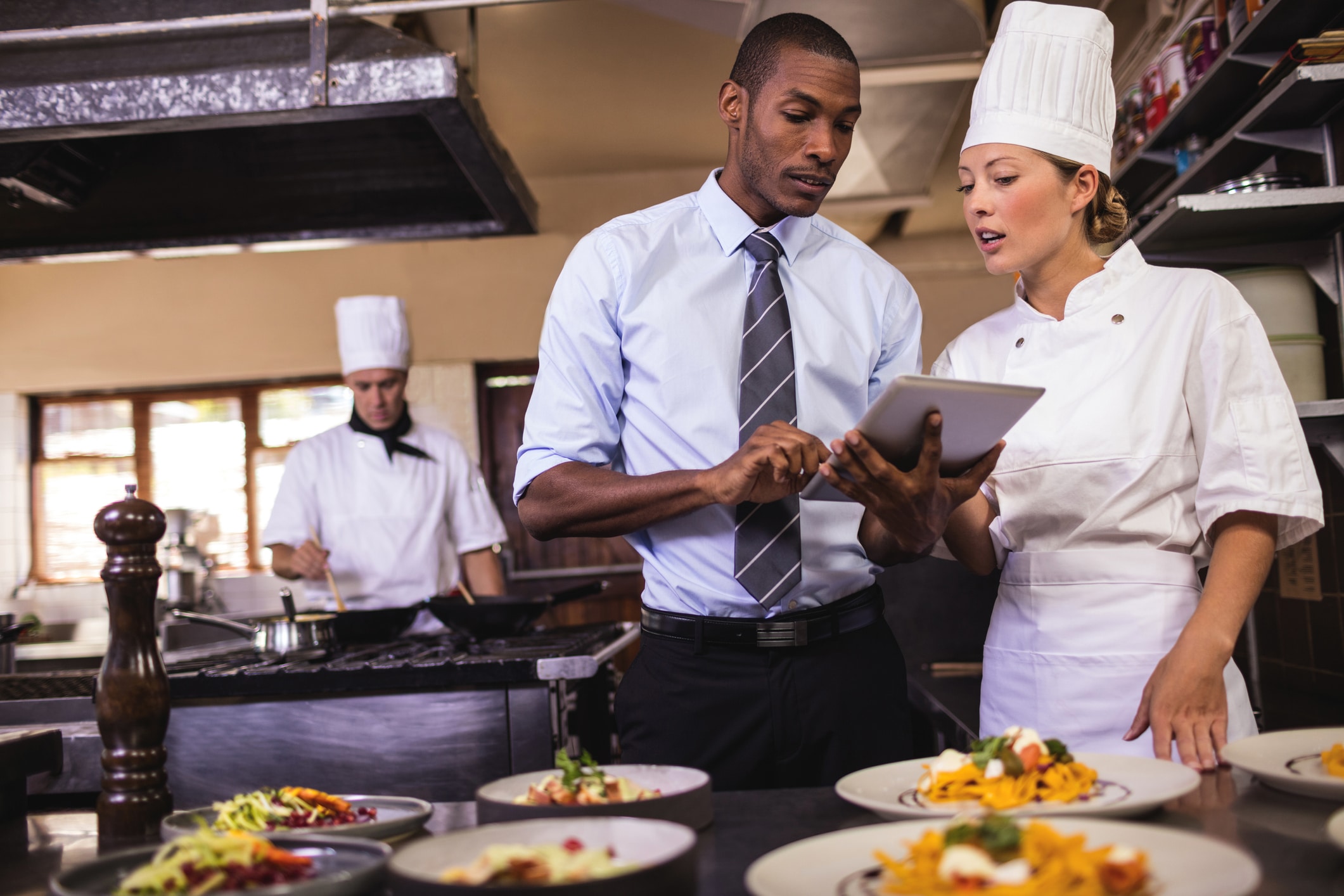 Un homme en costume et une chef regardent une tablette dans la cuisine d'un restaurant, tandis qu'un autre chef cuisine à l'arrière-plan. Des assiettes de plats sont disposées sur le plan de travail devant eux.