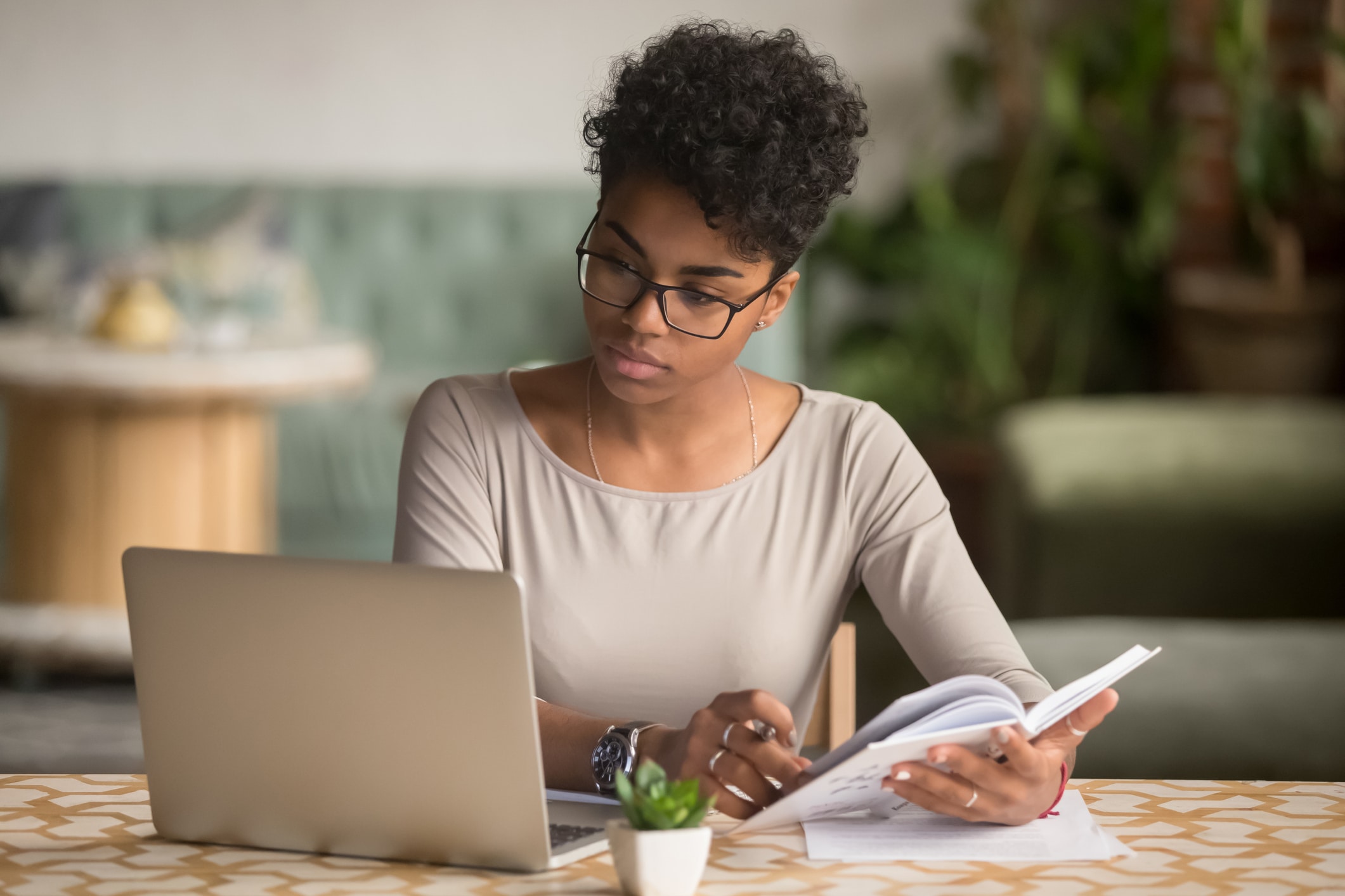 Une femme portant des lunettes est assise à une table ; elle regarde un ordinateur portable tout en tenant un cahier et un stylo, l'air concentrée et absorbée par son travail. On aperçoit des plantes et un canapé à l'arrière-plan.